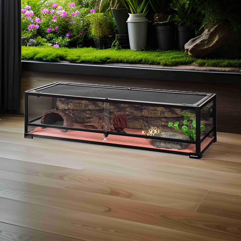Long glass reptile terrarium with mesh lid, artificial plants, rocks, and wood decor on hardwood floor